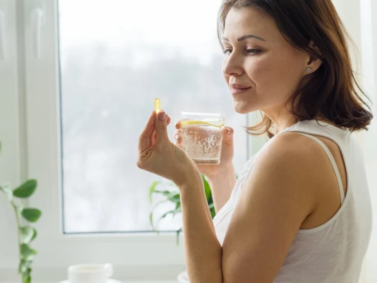 Woman looking at a softgel capsule while holding a glass of lemon-infused water