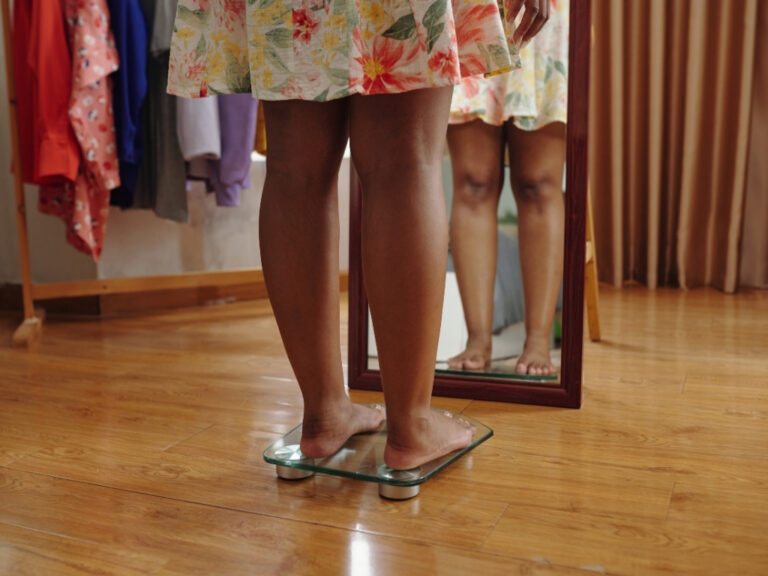 Woman in a floral dress checking her weight on a scale while in front of a full-body mirror