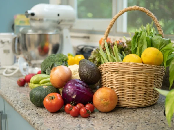 Basket of fresh vegetables and lemons on a kitchen counter, surrounded by tomatoes, onions, avocado, broccoli, and leafy greens