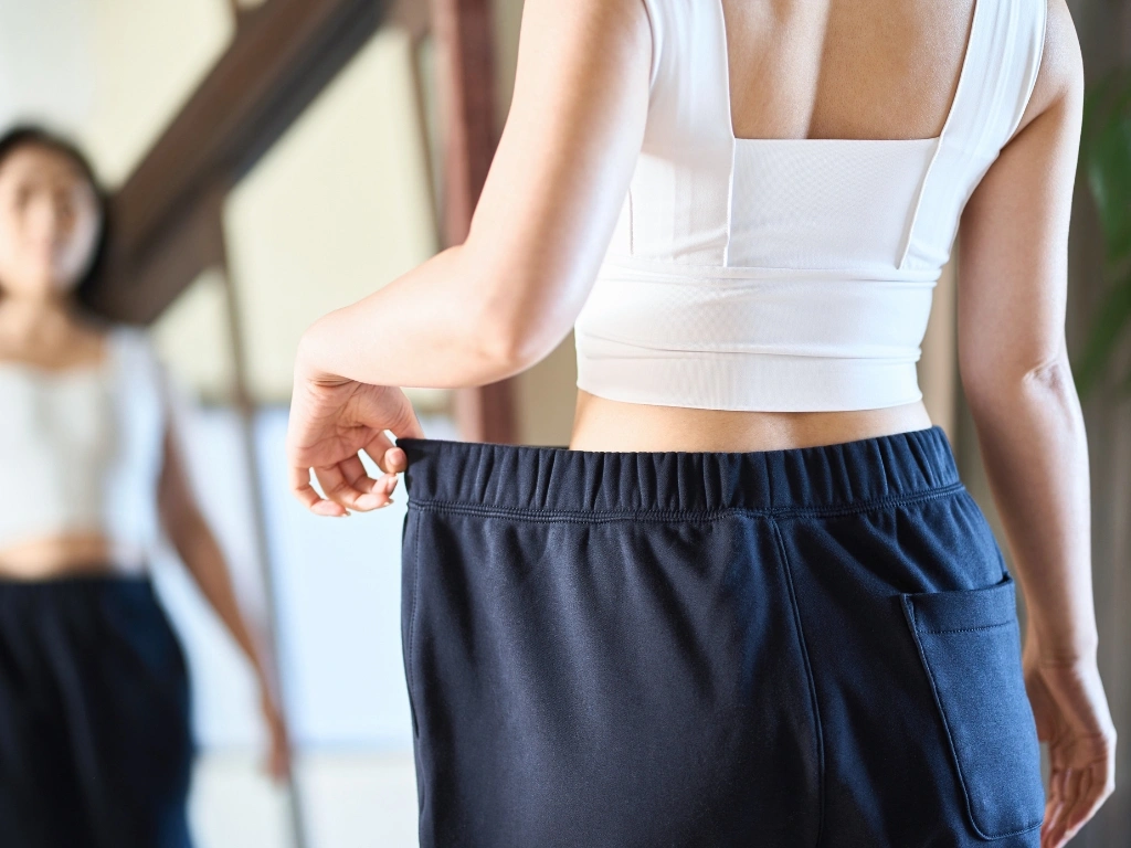 Woman wearing a white top holds out her loose pants in the mirror