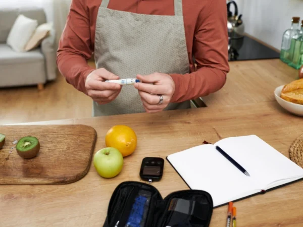 Man in an apron holding a blood glucose lancing device near fresh fruit and a logbook