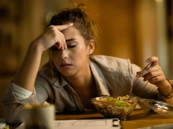 Woman holding her forehead in stress while gripping a fork in a bowl of salad with her other hand