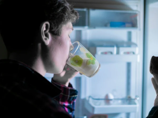 Man drinking from a glass and holding food in front of an open, glowing refrigerator at night