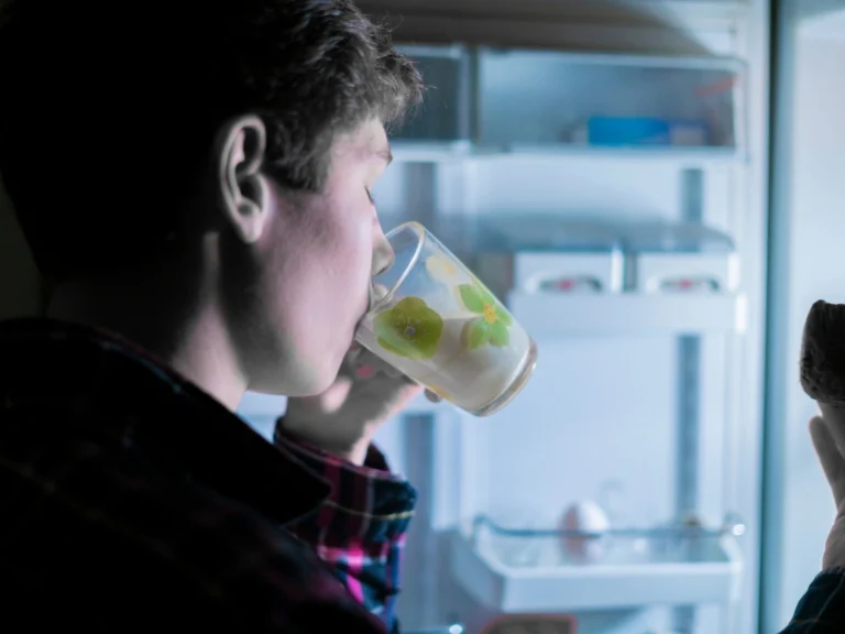 Man drinking from a glass and holding food in front of an open, glowing refrigerator at night