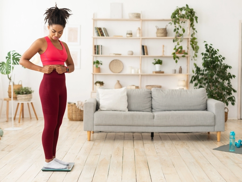 Sad Black Girl Touching Belly Standing On Scales At Home