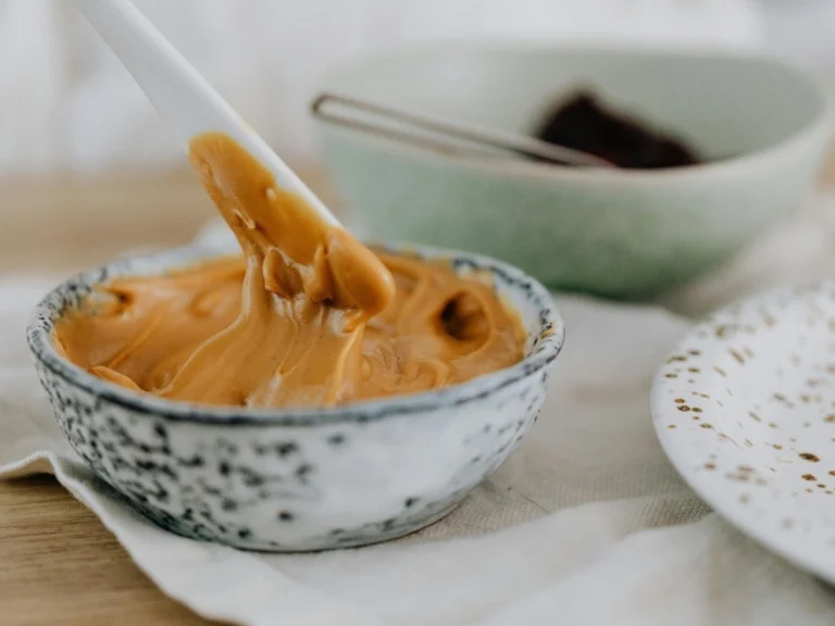 White plastic spoon stirring smooth, thinned-textured peanut butter in a small ceramic bowl