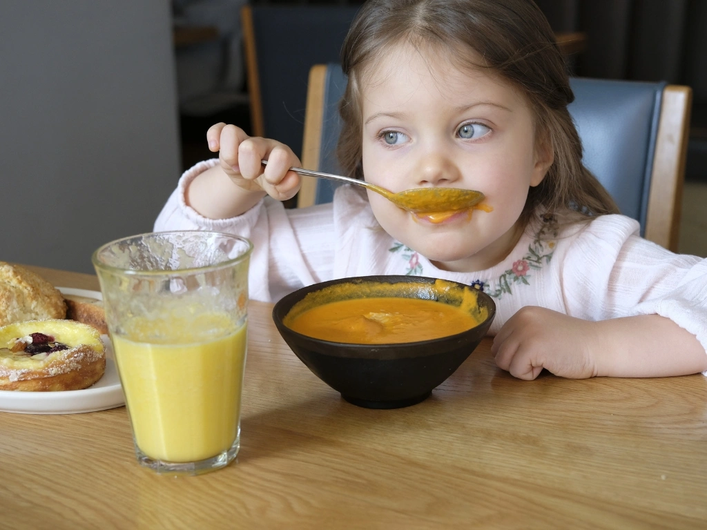 Toddler eating a bowl of tomato soup, with a glass of pineapple juice and a plate of sweet pastries also served on the table