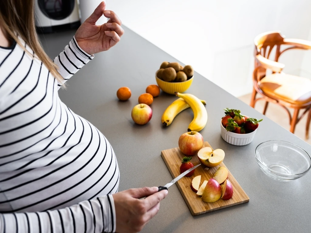 A pregnant woman slicing fresh fruits on a kitchen counter to prepare a healthy snack