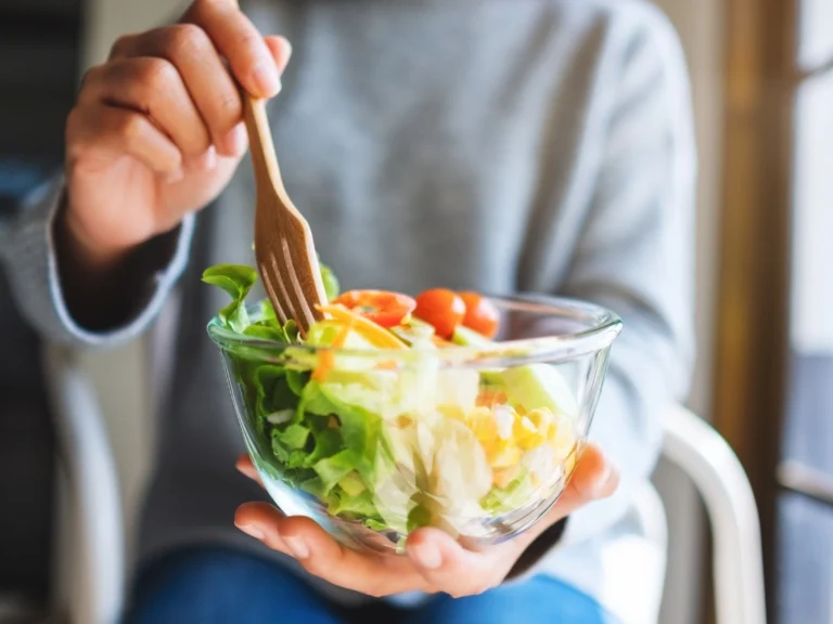 Woman in a long-sleeved top holding a bowl of vegetable salad with one hand and a wooden fork with the other