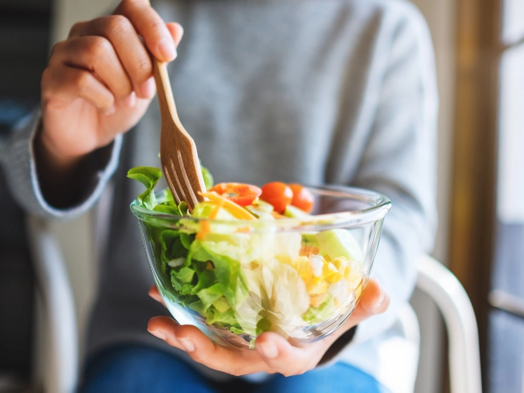 Woman in a long-sleeved top holding a bowl of vegetable salad with one hand and a wooden fork with the other