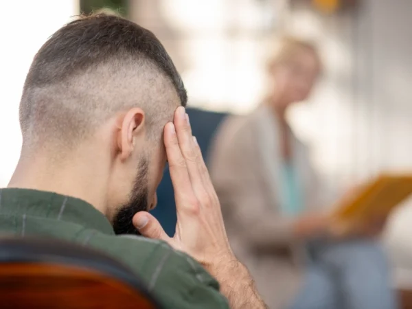 Man holding his temples due to a migraine, with a doctor looking in his direction in the background