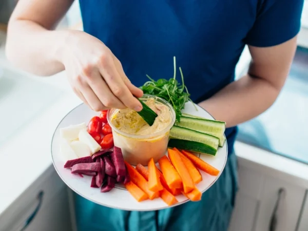 Person holding a plate of hummus and raw vegetables while dipping a cucumber slice
