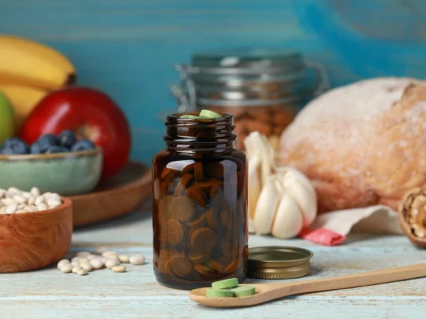 An amber medicine bottle filled with green supplement tablets and a wooden spoon holding some of the same tablets, surrounded by assorted food items on a wooden table