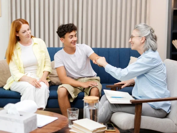 A smiling teenage boy bumping fists with a therapist after a successful therapy session, with a female guardian sitting next to him