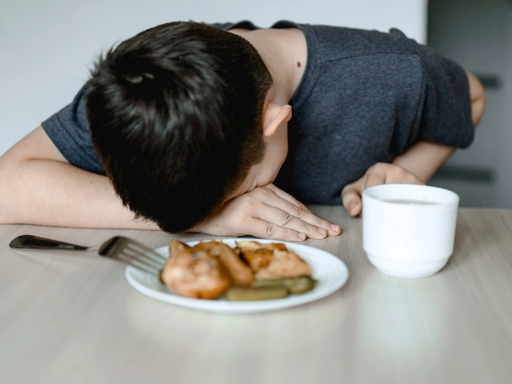 Man resting his head on a table next to a plate of chicken, pickles and a white mug