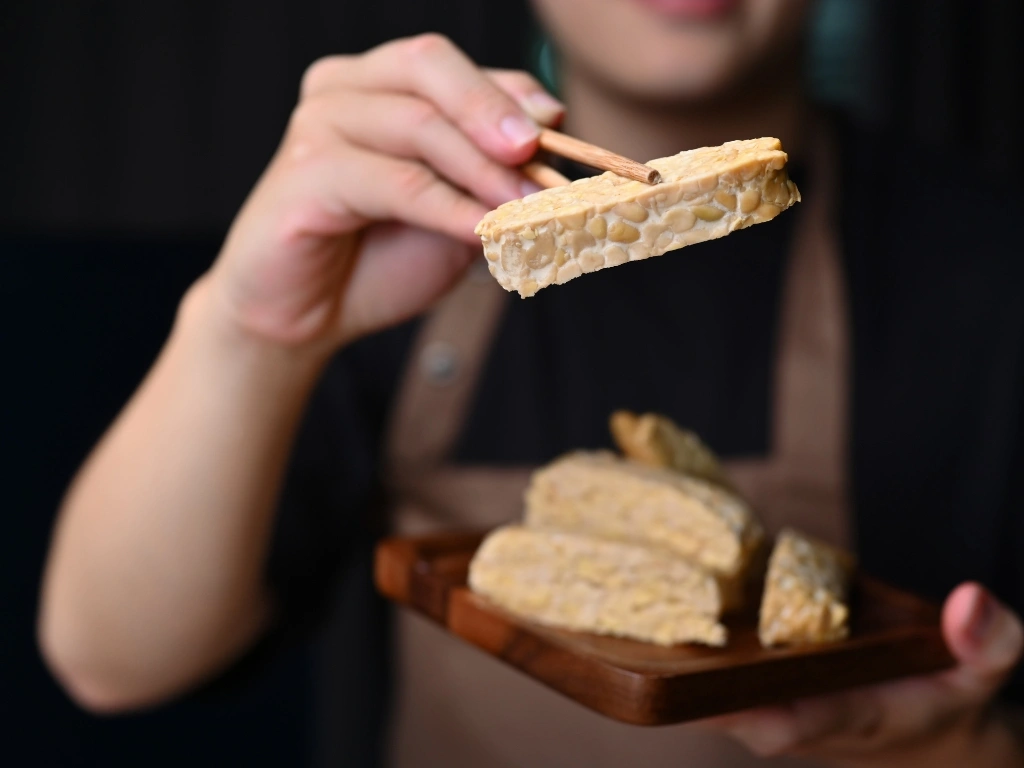 Person holding a slice of raw tempeh with chopsticks over a wooden plate of sliced tempeh