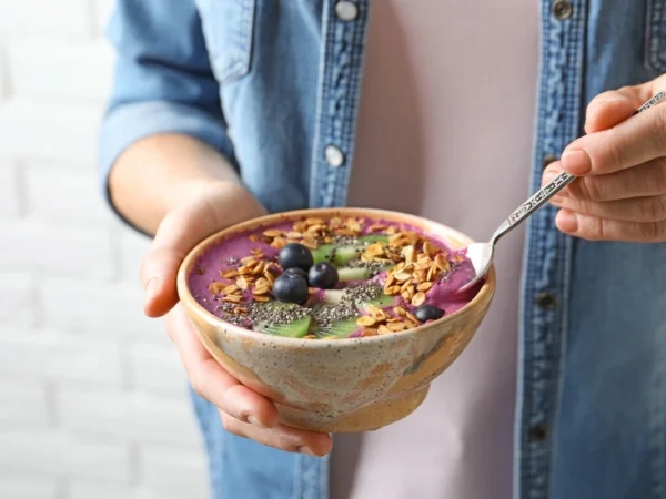 Person holding a ceramic bowl of vibrant purple smoothie topped with kiwi, granola, blueberries, and chia seeds