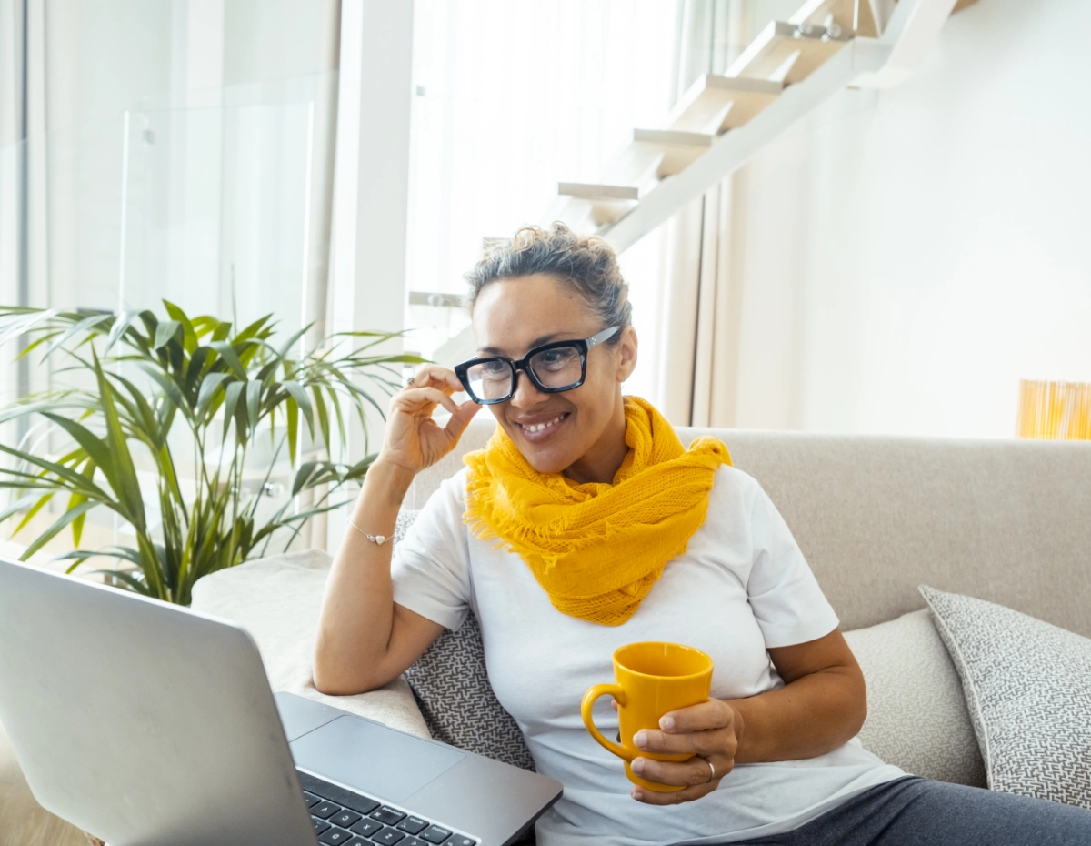 A smiling woman sits on a light-colored sofa, focusing on a laptop