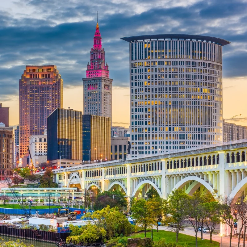 A cityscape of Cleveland at dusk, showing the downtown skyline with a prominent arched bridge over a river in the foreground
