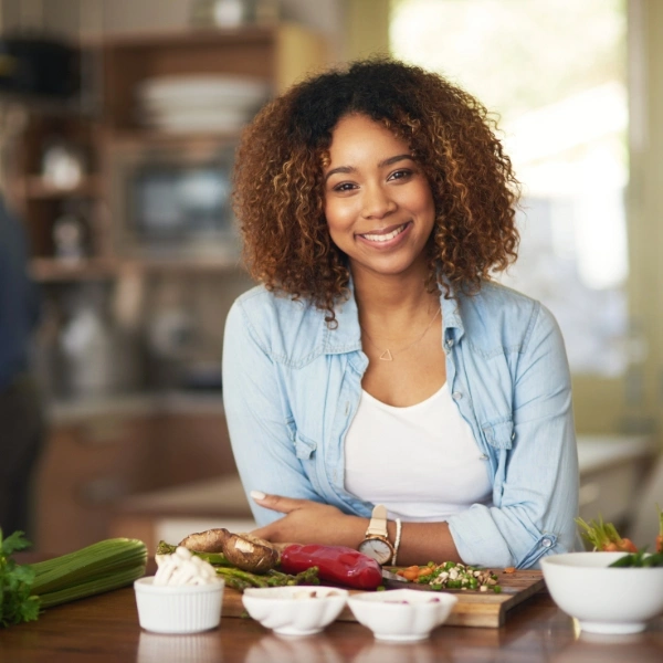 A smiling woman with curly brown hair wearing a light blue denim shirt, standing behind a kitchen counter with fresh vegetables and ingredients.