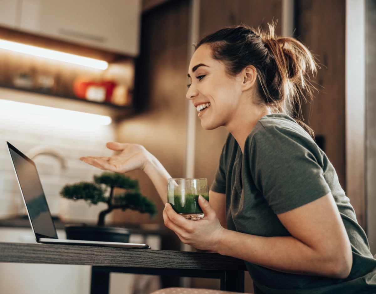 A smiling young woman holds a glass of green smoothie during a video call